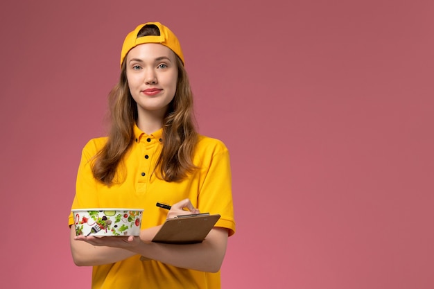 Vooraanzicht vrouwelijke koerier in geel uniform en cape met bezorgkom en blocnote schrijven op lichtroze muur service werknemer levering uniform baan bedrijf