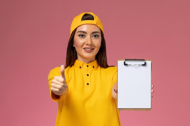 Vooraanzicht vrouwelijke koerier in geel uniform en cape met blocnote met glimlach op roze muur uniforme levering van de bedrijfsdienst