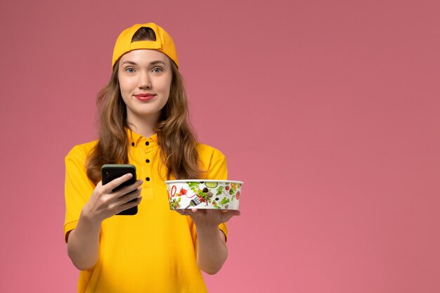 Vooraanzicht vrouwelijke koerier in geel uniform en cape met leveringskom en telefoon op roze muur