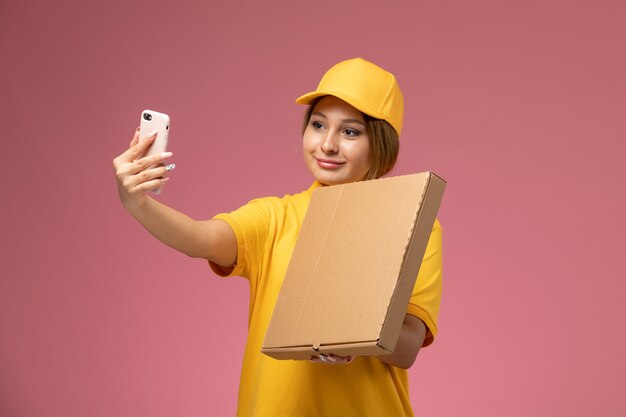 Vooraanzicht vrouwelijke koerier in gele uniform gele cape een selfie met eten op de roze achtergrond uniforme levering werkbaan