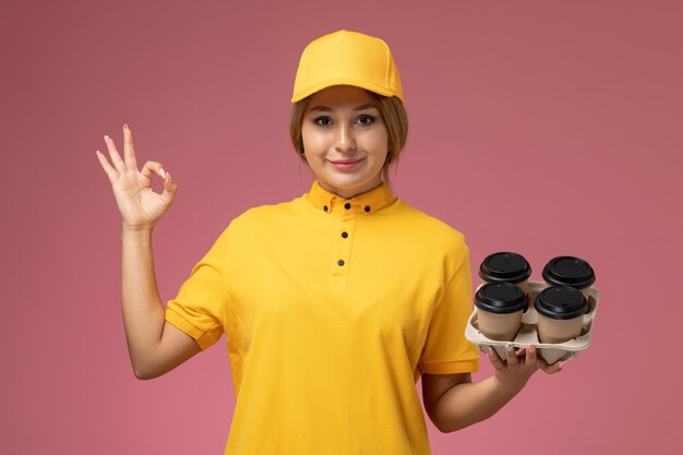 Vooraanzicht vrouwelijke koerier in gele uniform gele cape koffiekopjes met glimlach op roze achtergrond uniforme levering werk kleur baan te houden