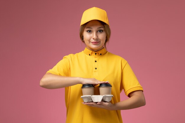 Vooraanzicht vrouwelijke koerier in gele uniform gele cape met bruine plastic koffiekopjes op de roze achtergrond uniforme levering werk kleur baan