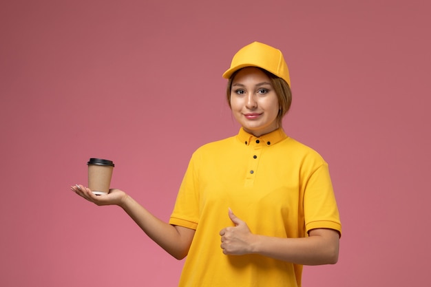 Vooraanzicht vrouwelijke koerier in gele uniform gele cape met plastic bruine koffiekopje met glimlach op de roze bureau uniforme levering vrouwelijke kleur