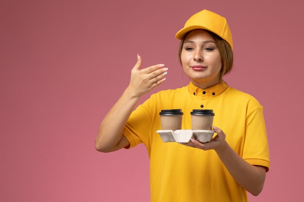 Vooraanzicht vrouwelijke koerier in gele uniform gele cape met plastic bruine koffiekopjes ruiken ze op het roze bureau uniforme levering vrouwelijke kleur