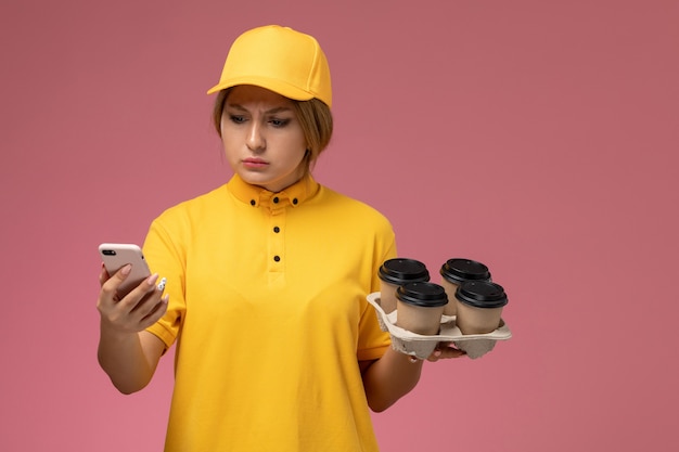 Vooraanzicht vrouwelijke koerier in gele uniform gele cape met plastic koffiekopjes en met behulp van telefoon op roze bureau uniforme levering werk kleur baan