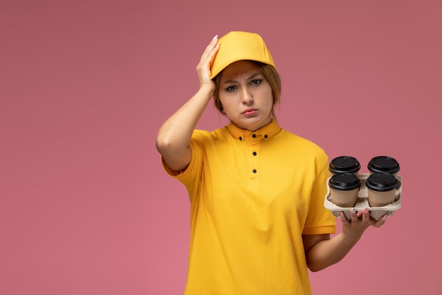 Vooraanzicht vrouwelijke koerier in gele uniform gele cape met plastic koffiekopjes met hoofdpijn op roze achtergrond uniforme levering werk kleur baan