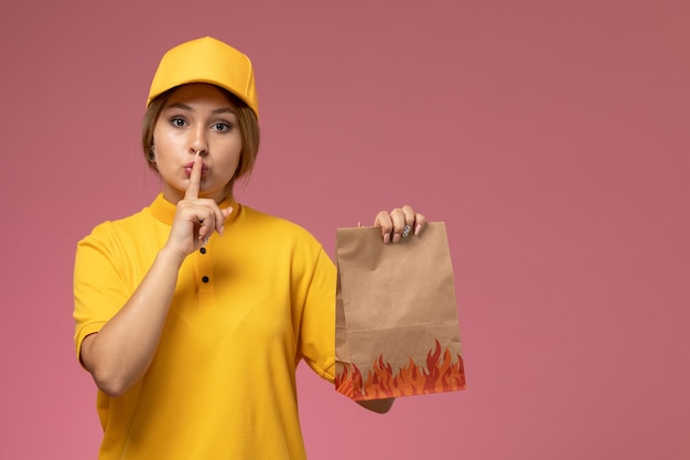 Vooraanzicht vrouwelijke koerier in gele uniform gele cape met voedselpakket met stilte teken op het roze bureau uniforme levering werk kleur baan