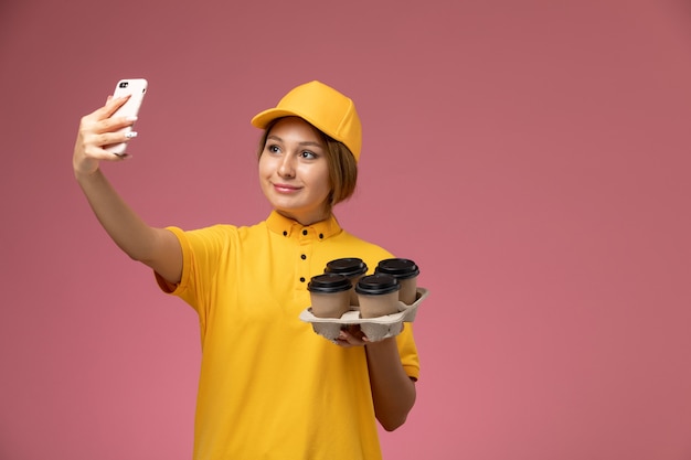 Vooraanzicht vrouwelijke koerier in gele uniform gele cape nemen een selfie met koffie op roze achtergrond uniforme levering werk kleur baan