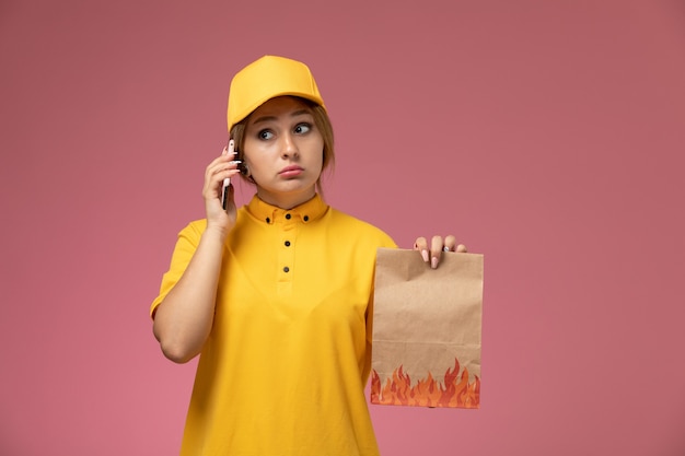 Vooraanzicht vrouwelijke koerier in gele uniform gele cape voedselpakket houden praten aan de telefoon op roze achtergrond uniforme levering werk kleur baan