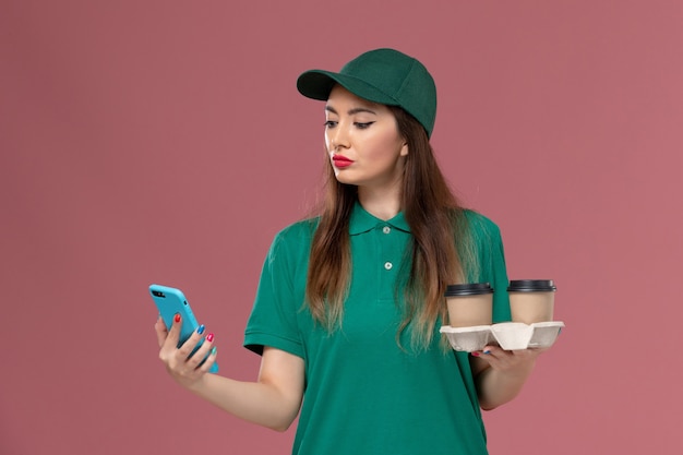 Vooraanzicht vrouwelijke koerier in groen uniform en cape met bezorging koffiekopjes en telefoon op roze muur service job uniforme levering