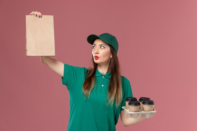 Vooraanzicht vrouwelijke koerier in groen uniform en cape met voedselpakket en bezorging koffiekopjes op roze bureau service uniforme levering baan werk