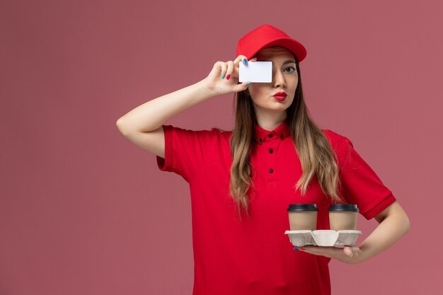 Vooraanzicht vrouwelijke koerier in rode uniforme levering koffiekopjes en plastic kaart op roze achtergrond service baan levering uniform