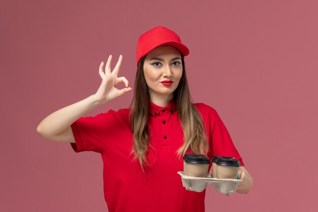 Vooraanzicht vrouwelijke koerier in rode uniforme levering koffiekopjes glimlachend op roze achtergrond werknemer baan service levering uniform