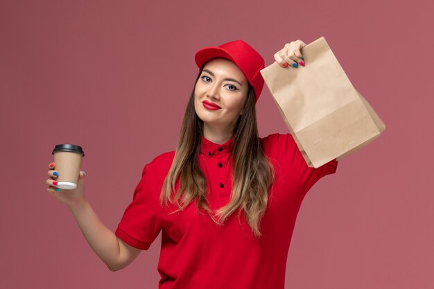 Vooraanzicht vrouwelijke koerier in rood uniform bedrijf levering koffiekopje en voedselpakket glimlachend op de roze achtergrond service levering uniform baan