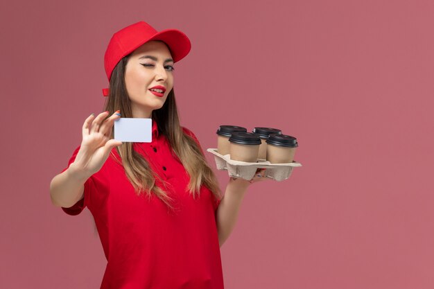 Vooraanzicht vrouwelijke koerier in rood uniform bedrijf levering koffiekopjes en kaart knipogen op roze achtergrond service levering bedrijf baan uniform