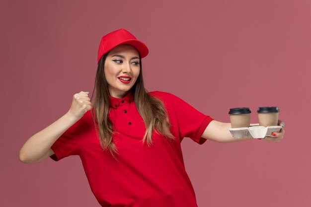 Vooraanzicht vrouwelijke koerier in rood uniform bedrijf levering koffiekopjes juichen op roze achtergrond werknemer baan service levering uniform