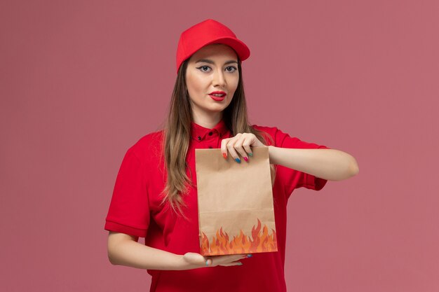 Vooraanzicht vrouwelijke koerier in rood uniform bedrijf papier voedselpakket op roze bureau werknemer service levering uniform bedrijf baan