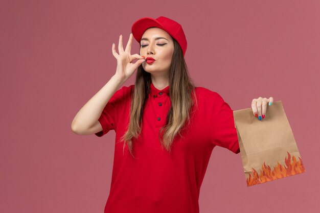 Vooraanzicht vrouwelijke koerier in rood uniform bedrijf papier voedselpakket smakelijk teken op roze achtergrond dienst baan levering uniform bedrijf tonen