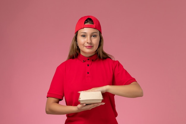 Vooraanzicht vrouwelijke koerier in rood uniform en cape bedrijf levering voedselpakket op lichtroze achtergrond werknemer uniforme bezorgdienst baan