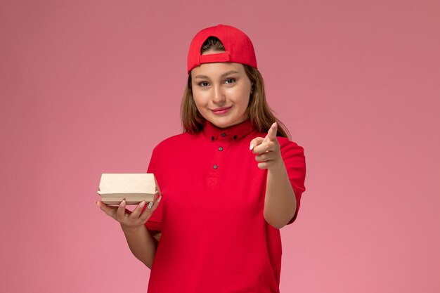 Vooraanzicht vrouwelijke koerier in rood uniform en cape bedrijf levering voedselpakket op roze achtergrond uniforme levering dienst bedrijf baan werk werknemer