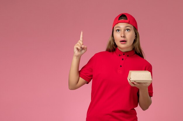 Vooraanzicht vrouwelijke koerier in rood uniform en cape die voedselpakket voor bezorging houden en denken aan roze muur, uniforme baanmedewerker van het bezorgbedrijf