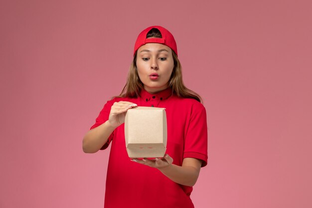 Vooraanzicht vrouwelijke koerier in rood uniform en cape die voedselpakket voor bezorging vasthoudt en opent op lichtroze muur, uniforme bezorgdienst baan werknemer bedrijf