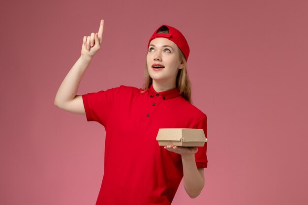 Vooraanzicht vrouwelijke koerier in rood uniform en cape die weinig voedselpakket voor bezorging op de lichtroze muur houdt, uniforme baan van bezorgbedrijf