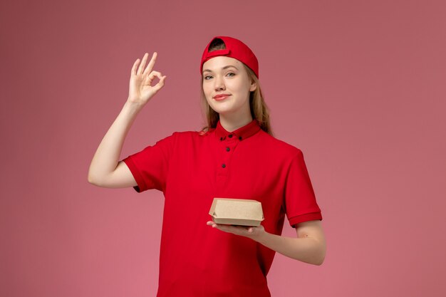 Vooraanzicht vrouwelijke koerier in rood uniform en cape die weinig voedselpakket voor bezorging op de roze muur houdt, uniforme werker van het bezorgbedrijf