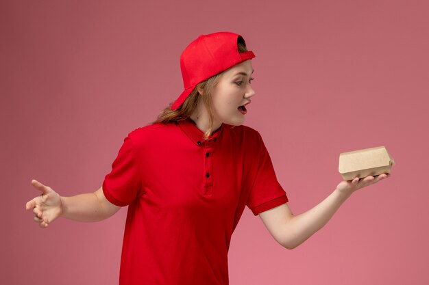 Vooraanzicht vrouwelijke koerier in rood uniform en cape die weinig voedselpakket voor bezorging op lichtroze muur houdt, uniforme werknemer van het bezorgbedrijf