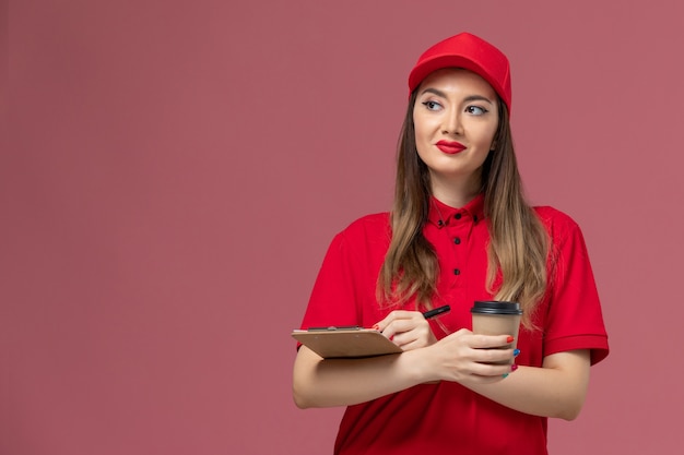 Vooraanzicht vrouwelijke koerier in rood uniform en cape levering koffiekopje met blocnote en pen op lichtroze achtergrond service levering uniform