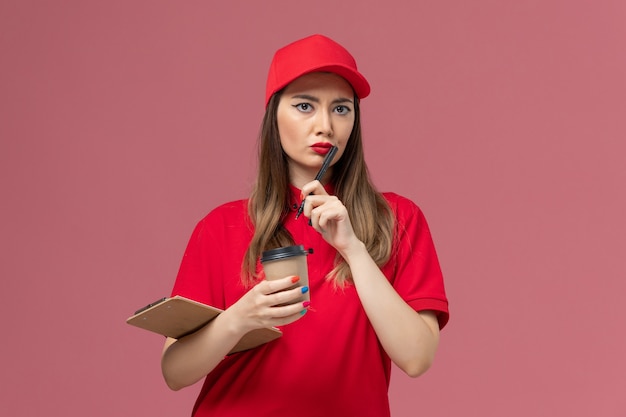 Vooraanzicht vrouwelijke koerier in rood uniform en cape met bezorging koffiekopje met blocnote en pen op de lichtroze achtergrond service job delivery uniform