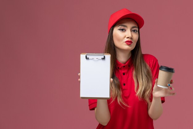 Vooraanzicht vrouwelijke koerier in rood uniform en cape met bezorging koffiekopje met blocnote op de lichtroze achtergrond service baan levering uniform