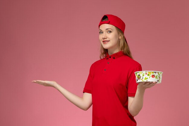 Vooraanzicht vrouwelijke koerier in rood uniform en cape met bezorgkom op de lichtroze muur, service uniform bezorgwerk