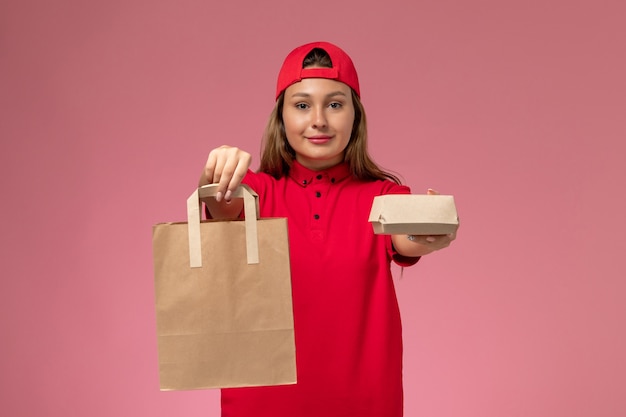 Vooraanzicht vrouwelijke koerier in rood uniform en cape met bezorgvoedselpakketten op de roze muur, uniforme bezorgdienstmedewerker