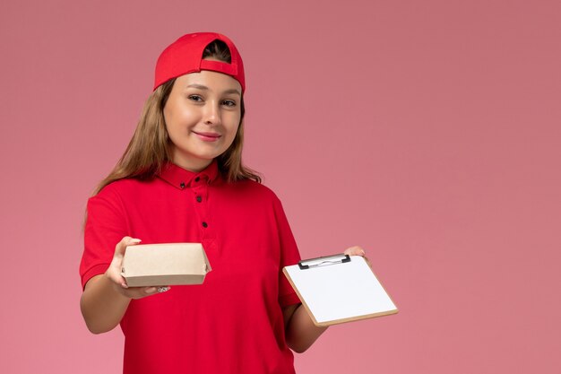 Vooraanzicht vrouwelijke koerier in rood uniform en cape met een klein pakket met voedsel voor bezorging en blocnote op de roze muur, een baan voor een uniforme bezorgdienst