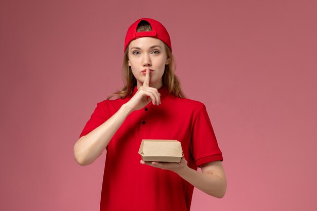 Vooraanzicht vrouwelijke koerier in rood uniform en cape met een klein pakket met voedsel voor bezorging op de roze muur, een uniforme baan voor bezorgdienst