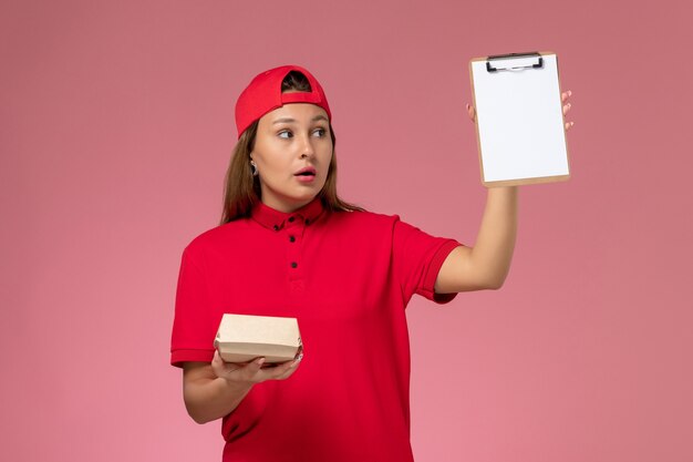 Vooraanzicht vrouwelijke koerier in rood uniform en cape met klein pakket met voedsel voor bezorging en blocnote op de roze muur, werknemer van uniforme bezorgdienst