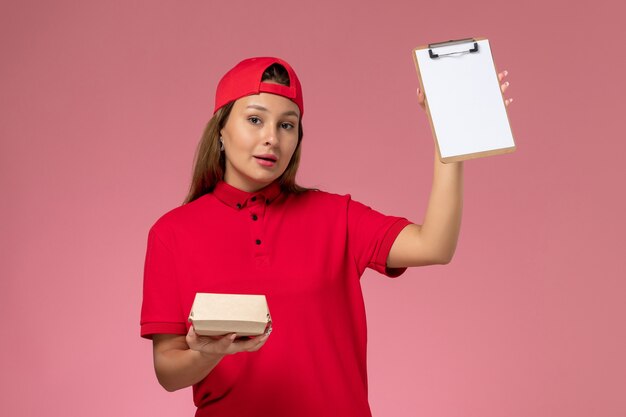 Vooraanzicht vrouwelijke koerier in rood uniform en cape met klein pakket met voedsel voor bezorging en blocnote op roze muur, baan voor uniform bezorgbedrijf