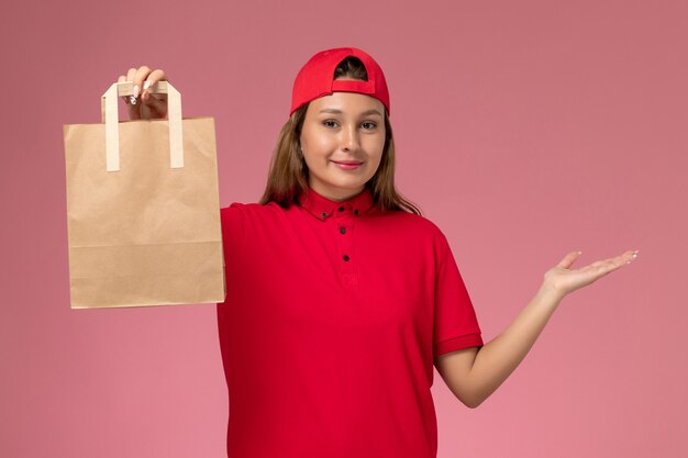 Vooraanzicht vrouwelijke koerier in rood uniform en cape met leveringsdocument pakket op de roze muur, baanuniform bezorgdienstmedewerker