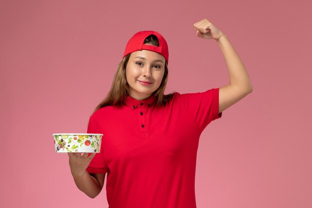 Vooraanzicht vrouwelijke koerier in rood uniform en cape met leveringskom buigen op roze muur, uniforme bezorgdienst voor baanarbeiders