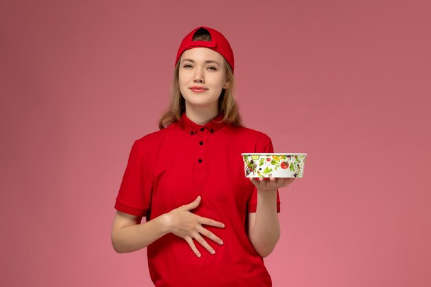 Vooraanzicht vrouwelijke koerier in rood uniform en cape met leveringskom op lichtroze muur, uniforme bezorger