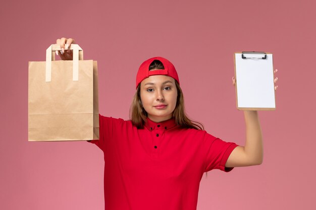 Vooraanzicht vrouwelijke koerier in rood uniform en cape met voedselpakket voor bezorging en blocnote op roze muur, uniforme bezorgdienst