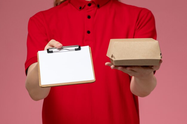 Vooraanzicht vrouwelijke koerier in rood uniform en cape met weinig bezorgvoedselpakket met blocnote op roze muur, service uniforme levering