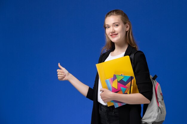 Vooraanzicht vrouwelijke student in wit overhemd en zwart jasje dragen rugzak met bestanden met voorbeeldenboeken op de blauwe muur college universitaire lessen