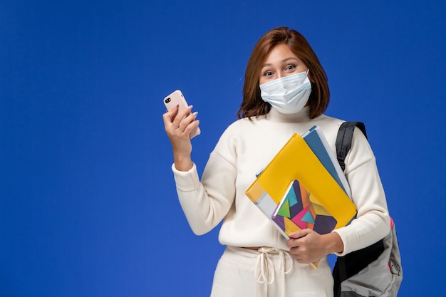 Vooraanzicht vrouwelijke student in witte trui dragen masker en rugzak met telefoon met koptelefoon op blauwe muur college universiteit boek lessen