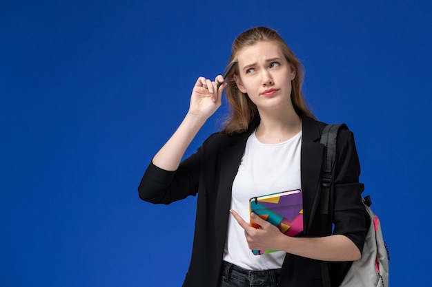 Vooraanzicht vrouwelijke student in zwart jasje dragen rugzak met pen en voorbeeldenboek op de blauwe muur college universitaire les