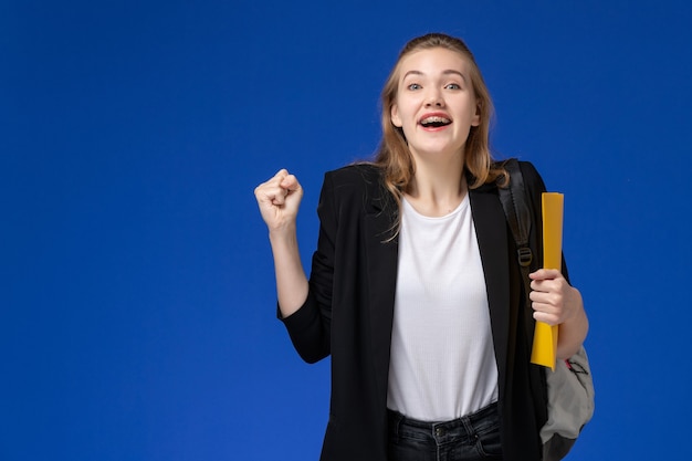 Vooraanzicht vrouwelijke student in zwarte jas draagt rugzak met gele bestanden en vreugde op de blauwe universiteit van de muurschool