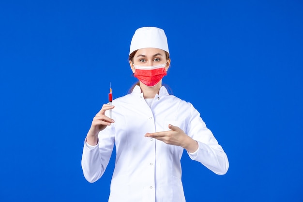 Vooraanzicht vrouwelijke verpleegster in wit medisch pak met rood masker en injectie in haar handen op blauw