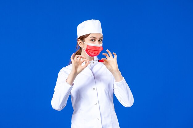 Vooraanzicht vrouwelijke verpleegster in wit medisch pak met rood masker en injectie in haar handen op blauw