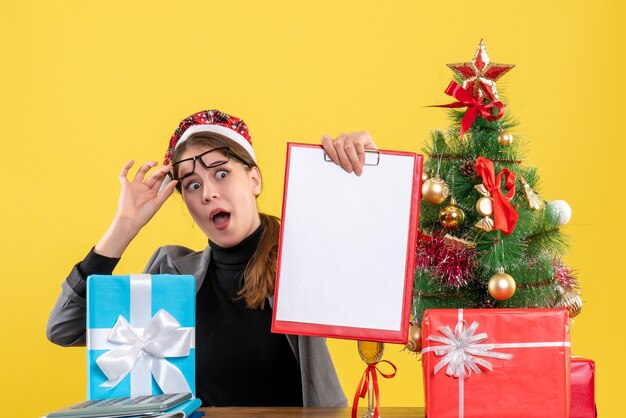 Vooraanzicht wide-eyed meisje met xmas hoed zittend aan de tafel opstijgen haar bril kerstboom en geschenken cocktail